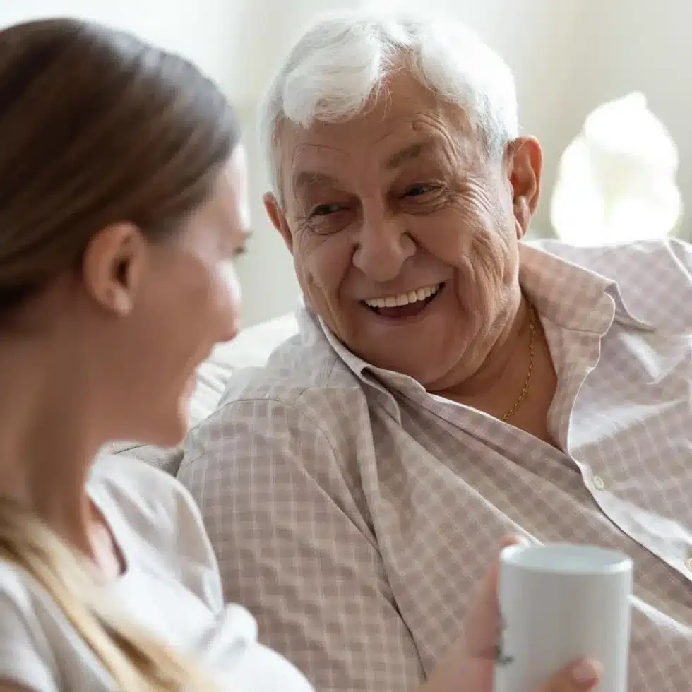 Elderly man with white hair smiling while chatting with a woman in a cozy home, holding a white mug alike a friendly moment.