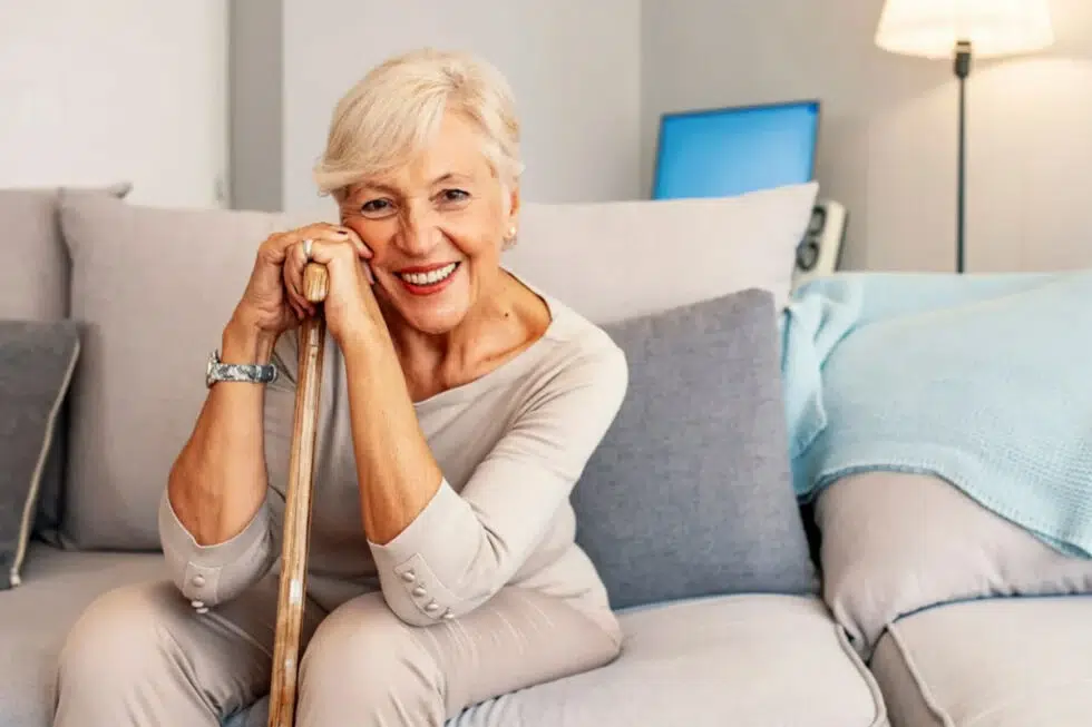 Smiling elderly woman on a living-room couch, resting her hands on a wooden walking cane.