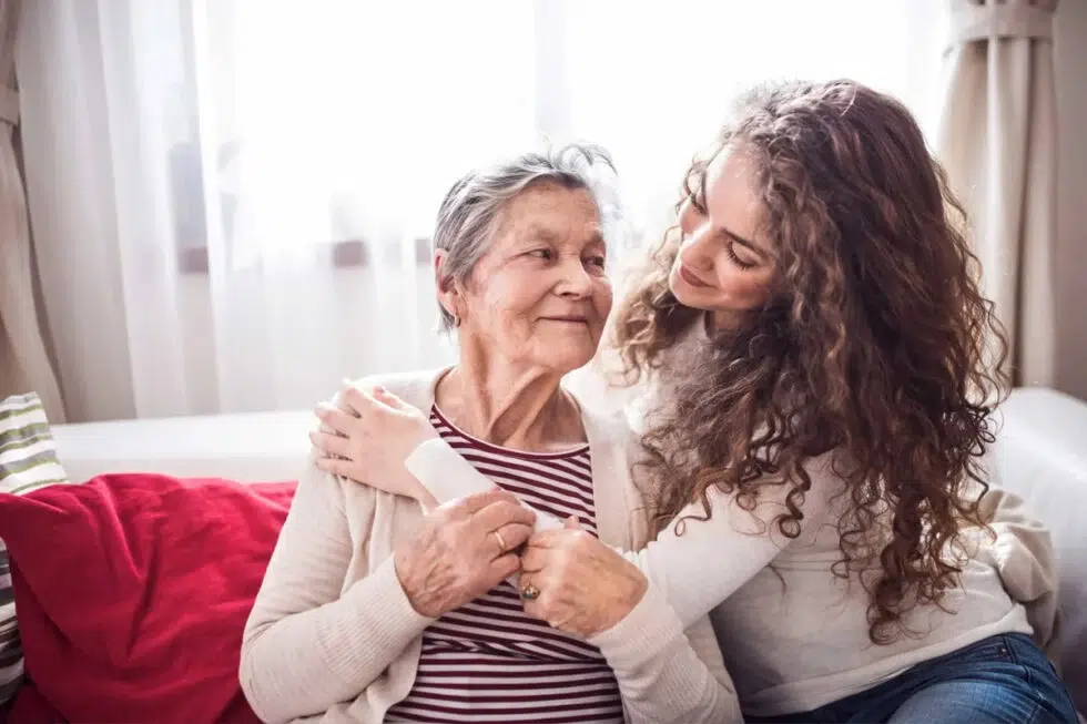 Young woman hugs an elderly woman on a bright living room couch on a sunny day.