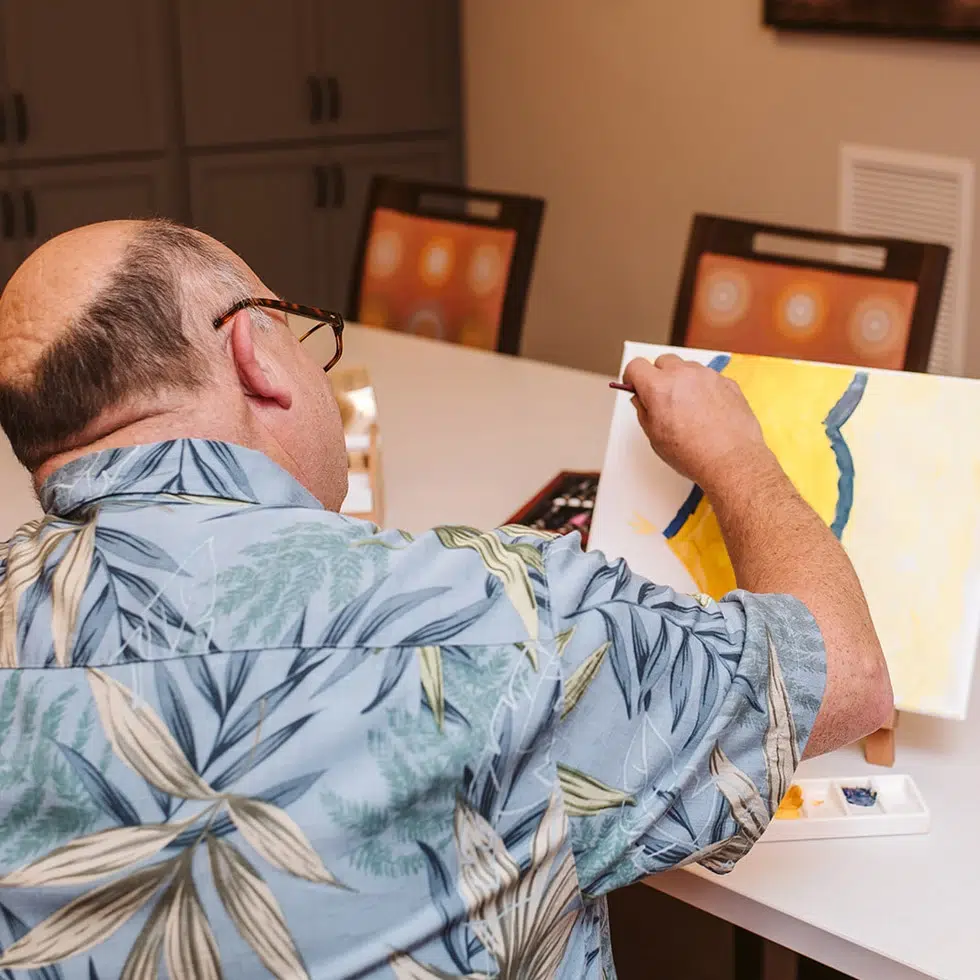 Back view of an artist in a Hawaiian shirt painting on a canvas at a table, watercolor palette nearby
