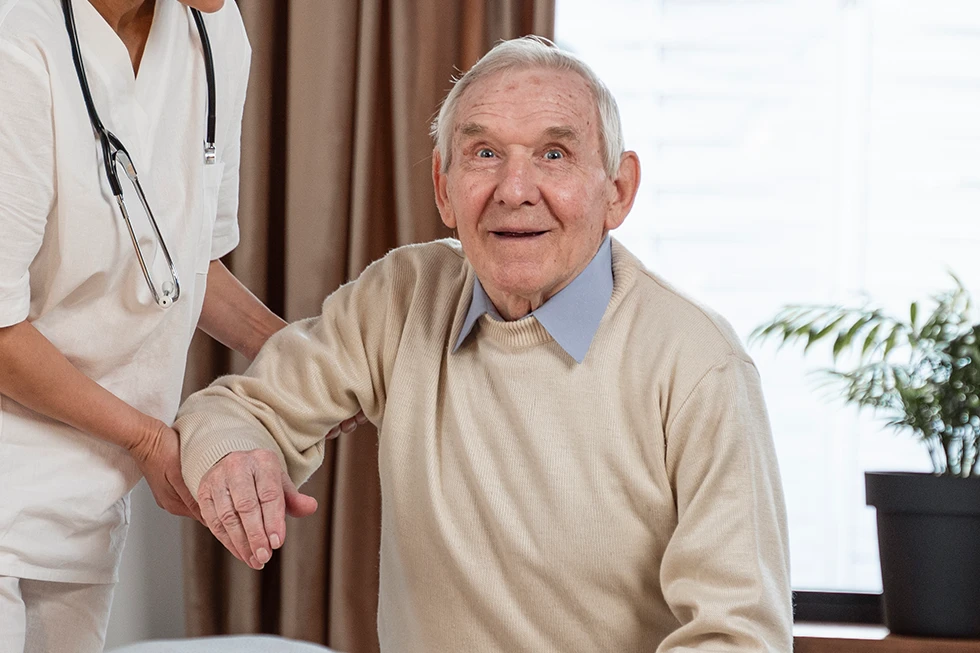 Caregiver helping an elderly man hold his arm during assistance, stethoscope visible around caregiver's neck